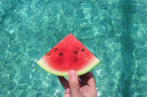 hand holding a slice of watermelon with blue swimming pool water in the background